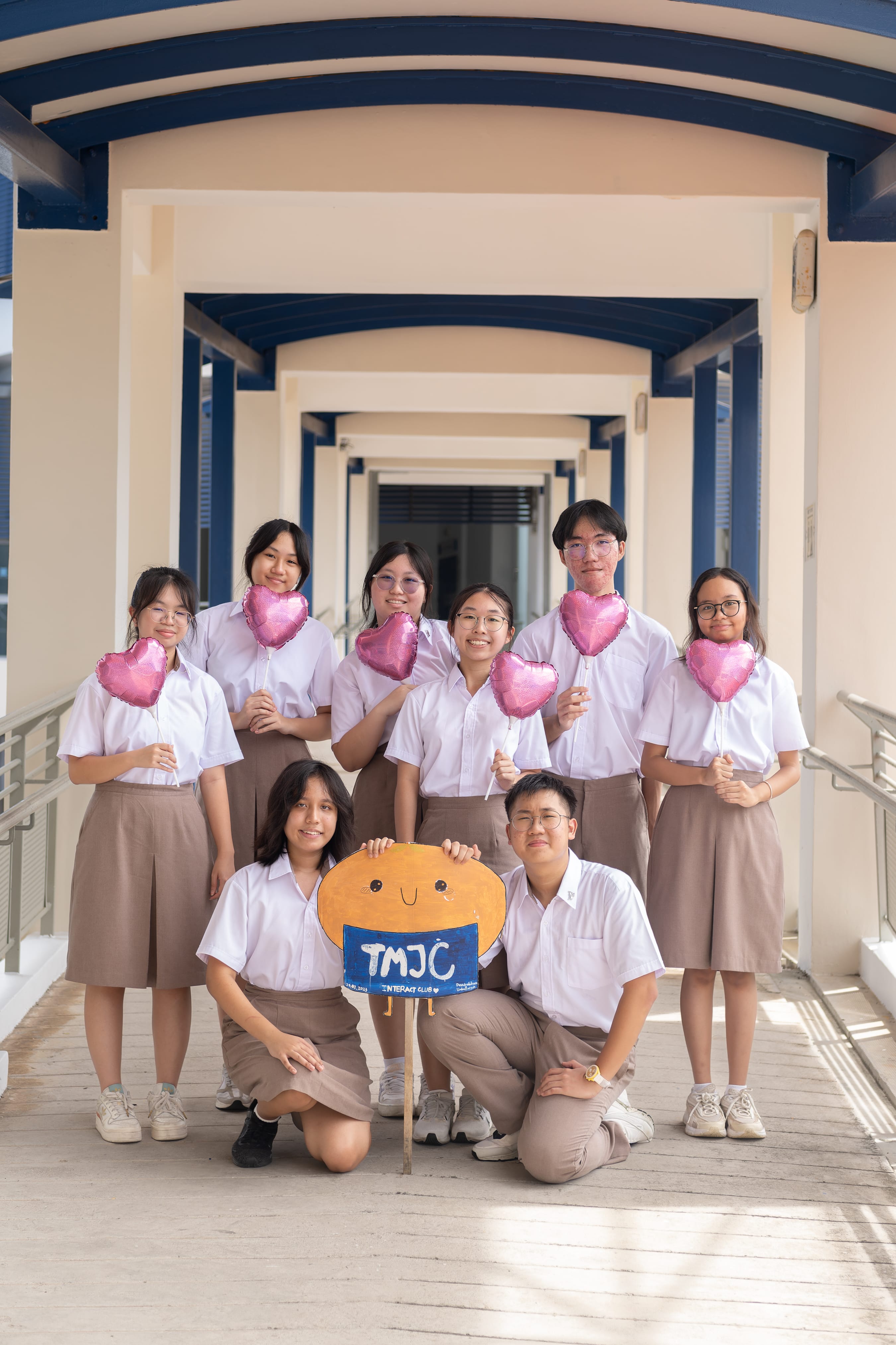Eight TMJC Interact Club students in white school uniforms pose cheerfully along a covered walkway, holding pink heart-shaped balloons. Two students in front kneel and hold a handmade sign featuring the club's mascot and the words "TMJC Interact Club".